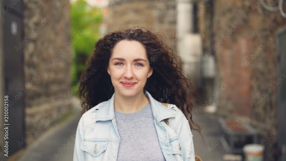 Close-up portrait of young lady happy tourist standing in the street against buildings in beautiful town and smiling looking at camera. City life and tourism concept. Close-up portrait of young lady happy tourist standing in the street against buildings in beautiful town and smiling looking at camera. City life and tourism concept.