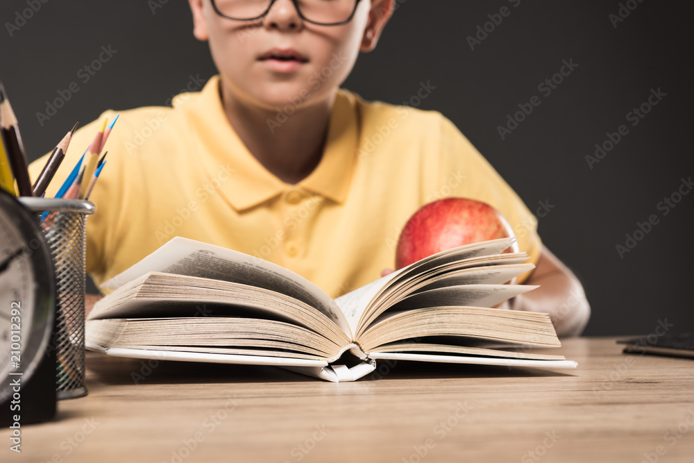 cropped image of schoolboy in eyeglasses eating apple and reading book at table with colour pencils and clock on grey background cropped image of schoolboy in eyeglasses eating apple and reading book at table with colour pencils and clock on grey background