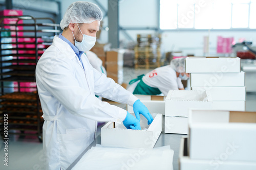 Side view of confectionery factory worker standing in white coat and preparing empty paper box. 