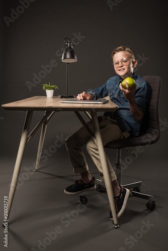 smiling little boy in eyeglasses holding pear and sitting at table with laptop, lamp and plant on grey background