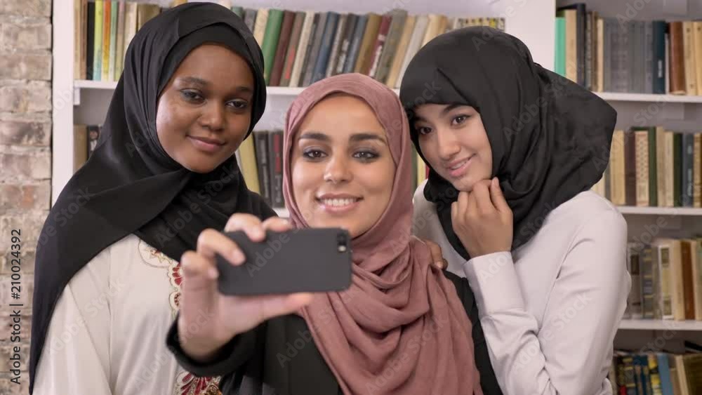 Three young beautiful muslim womens in hijab taking selfie in library and looking at phone after, smiling Three young beautiful muslim womens in hijab taking selfie in library and looking at phone after, smiling