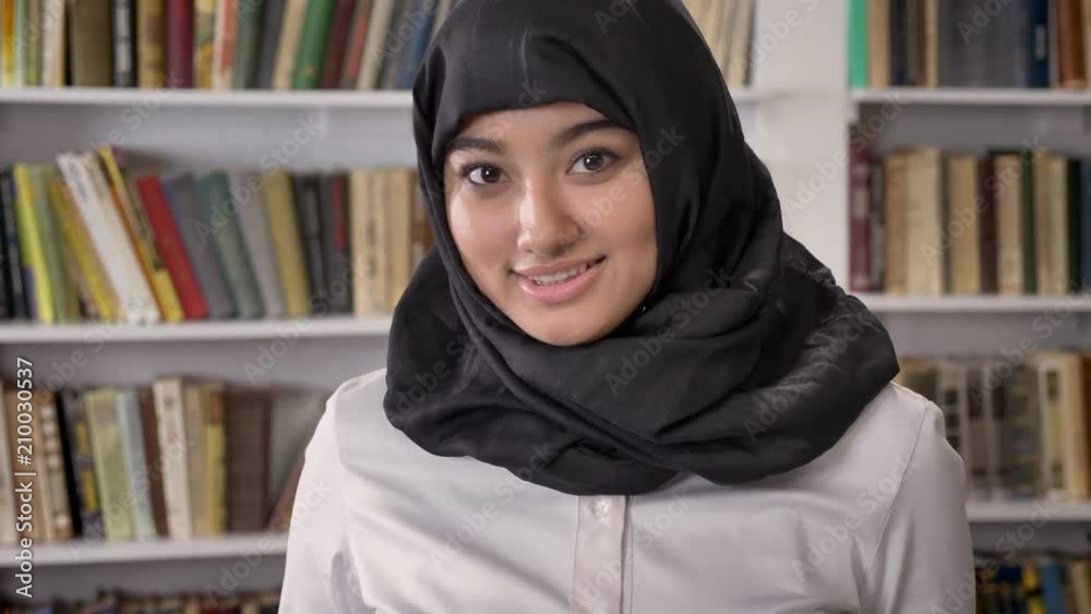 Young pretty muslim women in hijab looking into camera and smiling, standing in library, shelves with books background Young pretty muslim women in hijab looking into camera and smiling, standing in library, shelves with books background
