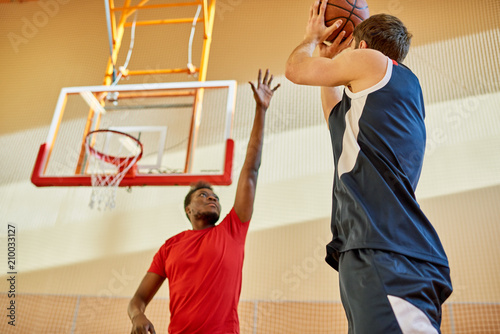 African-American man trying to stop contender from throwing ball into basket while playing basketball in gym. 