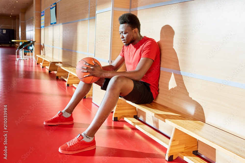 Obraz premium Attractive black basketball player holding ball and relaxing on bench during training in gym. 