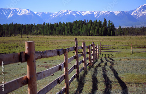Wooden fence with a shadow on a grass on a background of the mountains covered by a snow