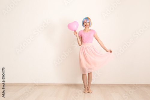 Beautiful young woman with colorful hair and balloon against light wall