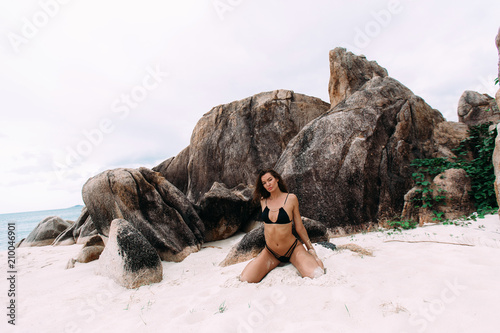 Sexy European model in expensive style swimsuit settles down on a white beach, sits on the sand against the backdrop of sharpened stones, feels the warm wind, the sound of water. photo