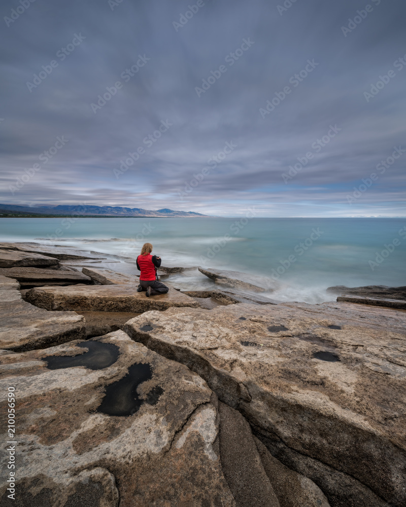 Red-Haired Girl-Photographer In Red Waistcoat On The Stony Shore Of Turquoise Sea Takes Pictures Of The Dawn. Girl And The Sea. Female Photographer In Red Clothes At Work, Long Exposure. Kyrgystan
