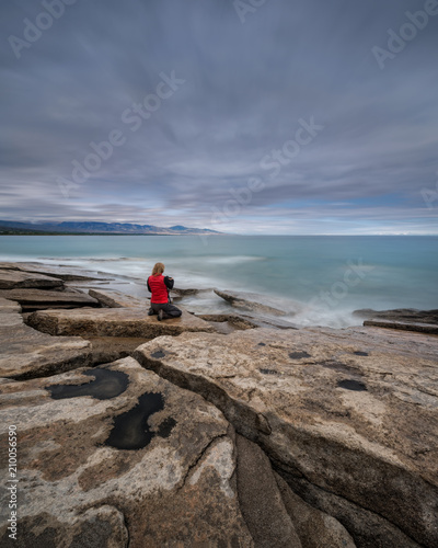 Obraz na płótnie Red-Haired Girl-Photographer In Red Waistcoat On The Stony Shore Of Turquoise Sea Takes Pictures Of The Dawn. Girl And The Sea. Female Photographer In Red Clothes At Work, Long Exposure. Kyrgystan w salonie
