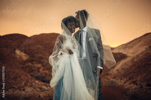 Newlyweds stand under bridal veil, smile and kiss in canyon at sunset. photo