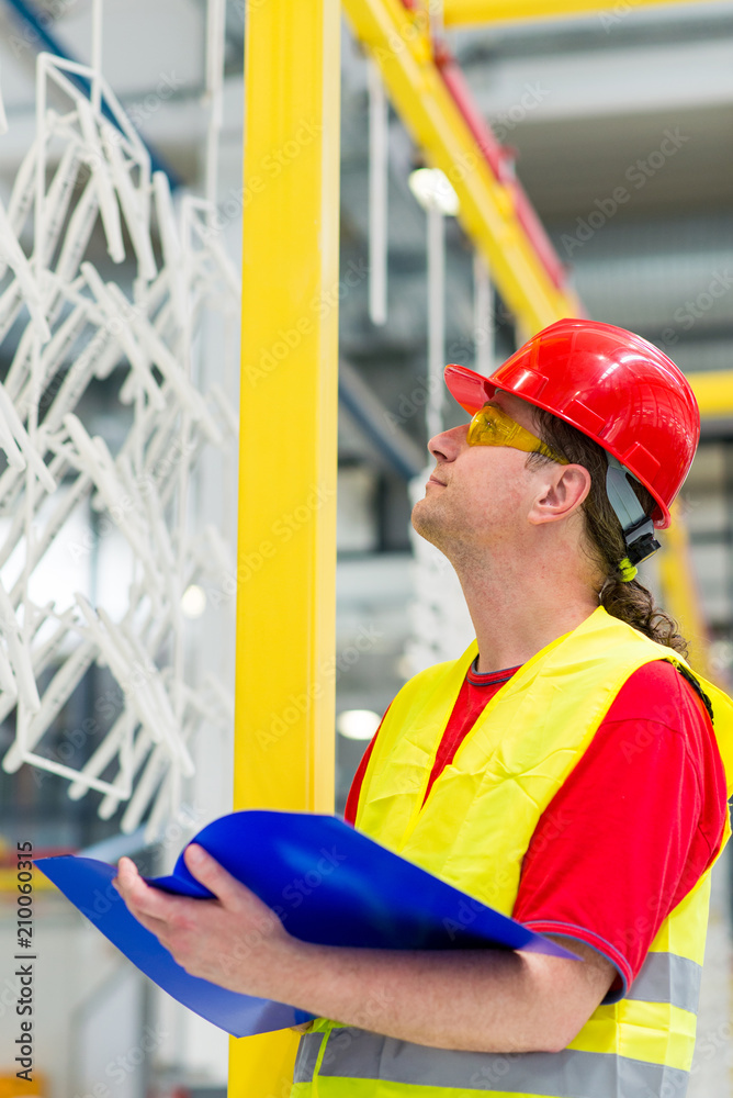 Factory supervisor in yellow reflective west with red helmet. Factory engineer checking production line holding blue folder with documents Factory supervisor in yellow reflective west with red helmet. Factory engineer checking production line holding blue folder with documents