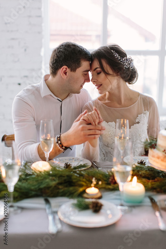 Bride and groom posing at the decorated banquet table in the white hall. Hand in hands. Enjoy a moment of happiness and love. A series of photos in my portfolio. Wedding flowers on a table