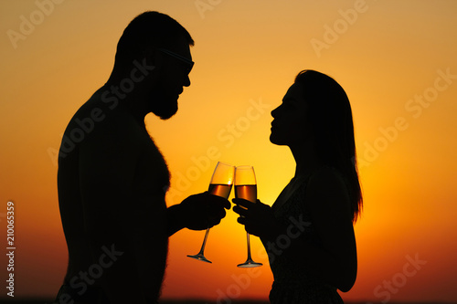 happy couple enjoying a glass of wine or champagne, silhouette of couple in love drinking wine from wineglasses during romantic dinner at sunset on the beach, sunset background