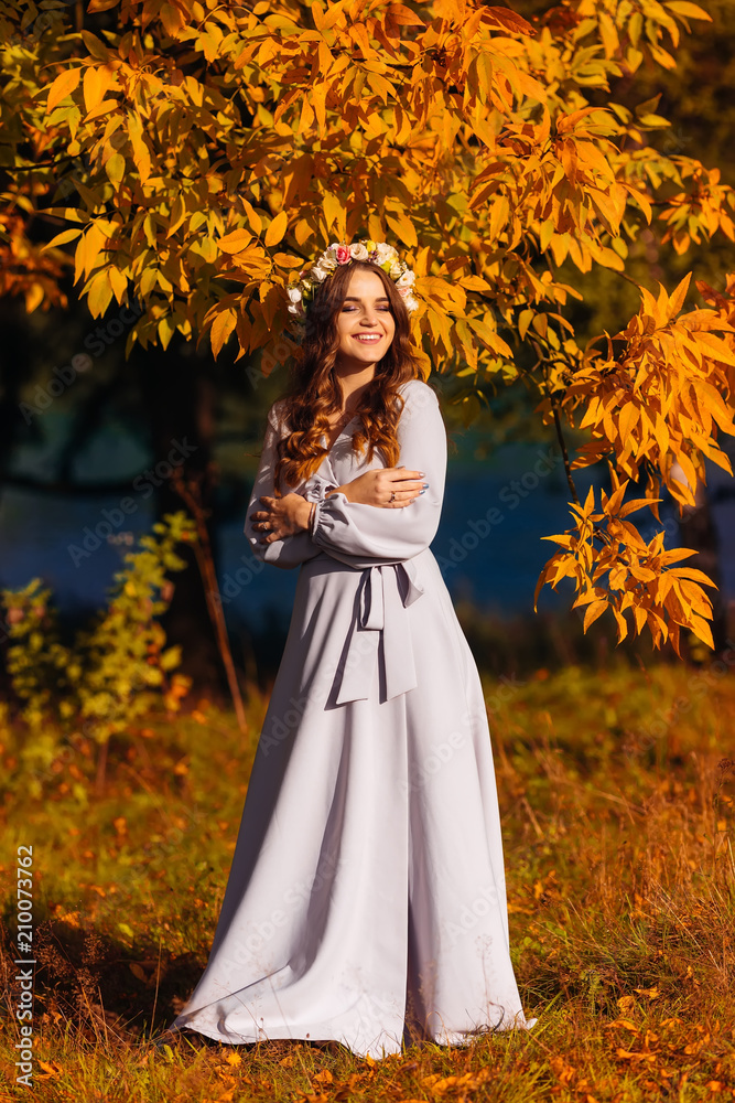 a woman in a long white dress closed her eyes and stands on the background of the lake and autumn trees