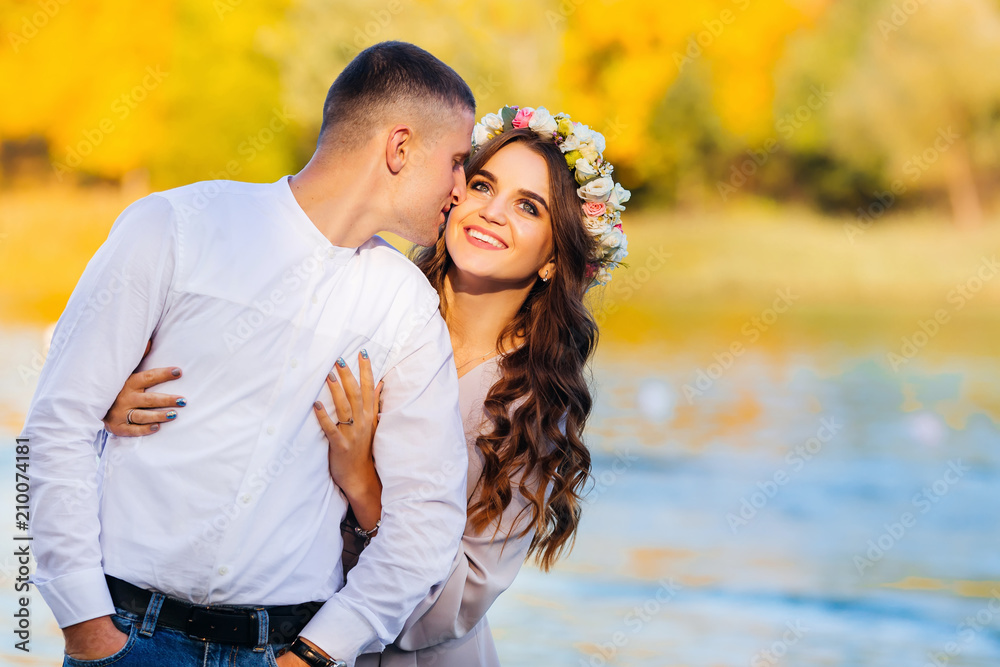 Fototapeta premium A man kisses his wife in a cheek and she smiles genuinely against the background of the lake and the trees.