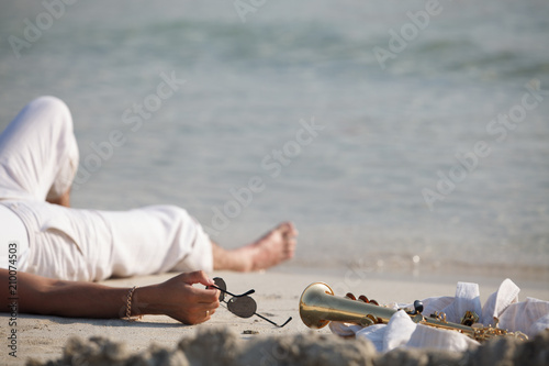 Мужчина лежит на песке и держит очки в руках. Саксофон на песке.