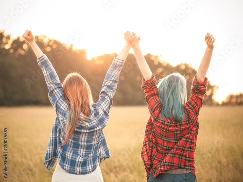 Two young women are standing on the background of sunset and rise hands up. Best friends