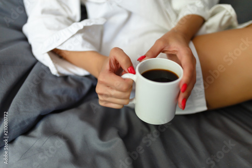 Slim, perfect and beautiful woman drinking her coffee while sitting on bed. Sunny Lazy morning in bed. Cropped image of erotic lying on the bed of a girl in the bedroom. Top view.