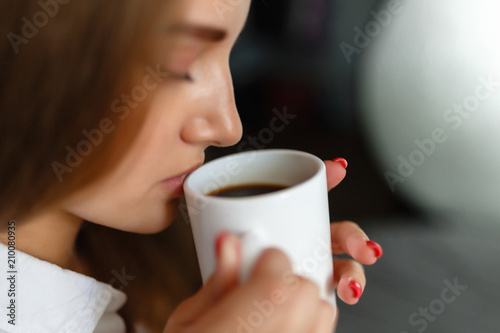 Close-up of the lips of a girl drinking coffee while sitting on the bed. Sunny lazy morning in bed. White mug with hot coffee in the hands of a young girl. Breakfast in bed.