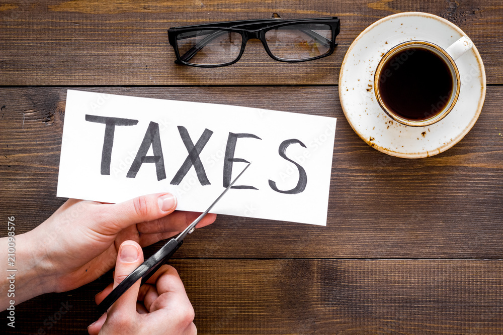 Tax reduce concept. Hands with sciccors cut paper with word Taxes on dark wooden background top view copy space Tax reduce concept. Hands with sciccors cut paper with word Taxes on dark wooden background top view copy space