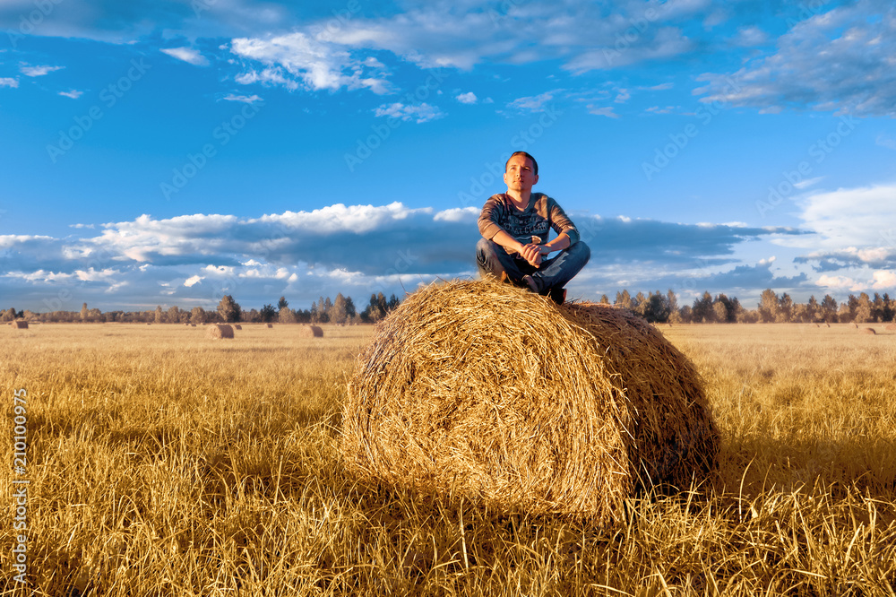 A young man sitting on a haystack, against the blue sky with clouds A young man sitting on a haystack, against the blue sky with clouds