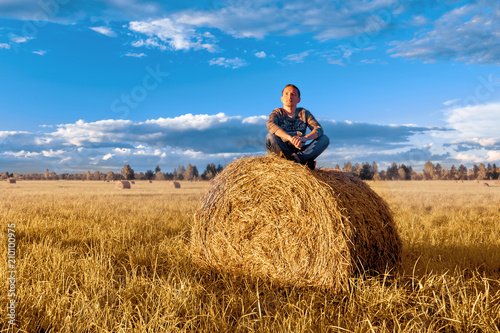 A young man sitting on a haystack, against the blue sky with clouds