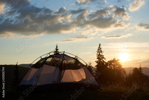 The tent stands on top of a mountain at dawn under the sky with rare clouds and a morning sun on a blurred background of mountains, hills and trees. The concept of healthy recreation in the mountains