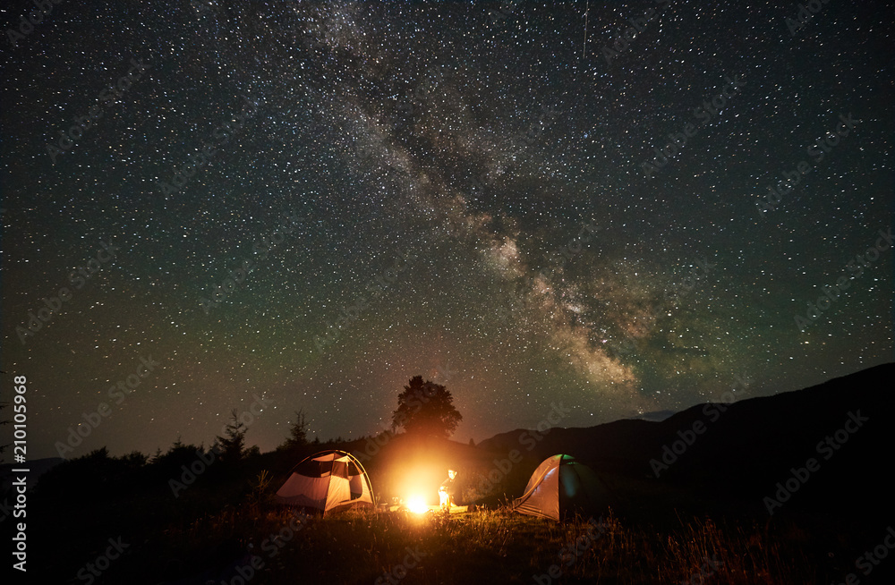 Woman camper resting at night camping in mountains under amazing night sky full of stars and milky way. Girl hiker sitting beside campfire and two tents. Concept of active lifestyle. Astrophotography