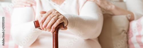 Close-up of an elderly woman's hands on a cane. Blurred background with upper torso of the senior with caretaker's arms around her shoulders. Panorama.