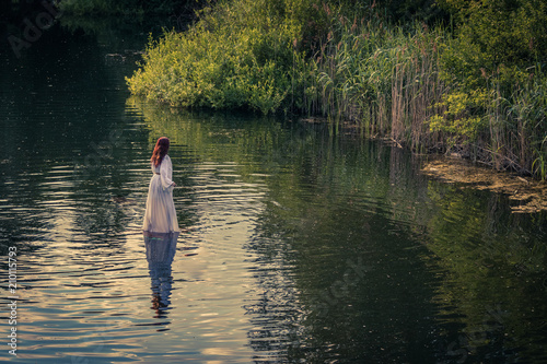 Red long hair Nymph woman in a white dress walking on the water surrounded by reeds trees and grasses 
