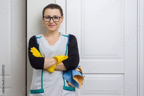 Professional cleaning service. Woman housemaid in yellow rubber gloves with detergents smiling against white doors photo