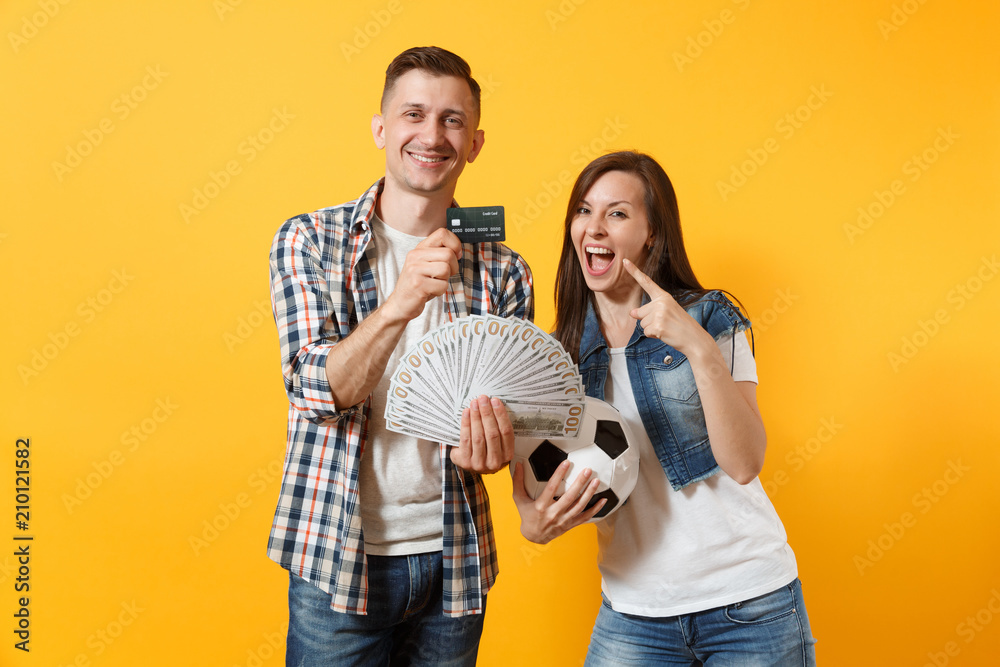 Fotka „Young win couple, woman man, football fans holding bundle of dollars  money, credit card, soccer ball, cheer up support team isolated on yellow  background. Sport bet, ardor family lifestyle concept.“ ze