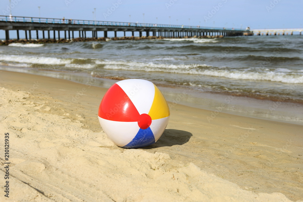 Obraz premium Colorful beach ball on sand and waving sea. Holiday beach ball near Baltic sea and Palanga bridge in horizon.