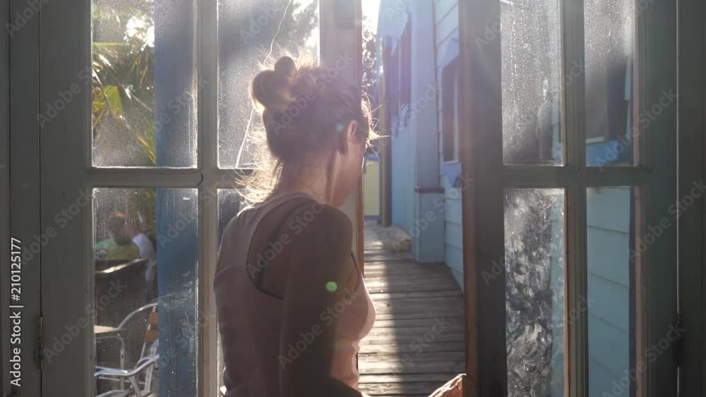 Woman goes away shutting the transparent glass door of wooden house in sun rays
