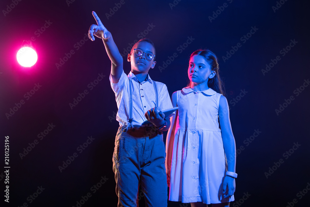 Fototapeta premium Modern technology. Unsmiling afro-american boy holding a tablet and a girl standing near him