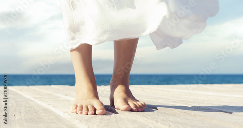 Beautiful and healthy feet of a young girl in white dress on a wooden pier. Vacation, resort and traveling concept.