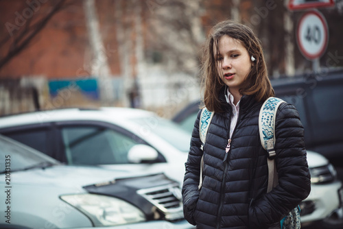 Schoolgirl with backpack walks in depressed state along city street in cloudy weather in rain. Young girl with headphones with wet hair in faded tones is outdoor with copy space. Melancholic teenager. © Daniil