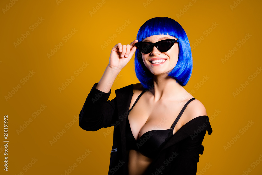 Portrait of positive toothy girl having fun good mood modern eyeglasses big boobs in bright blue wig holding eyelet with hand looking at camera isolated on yellow background