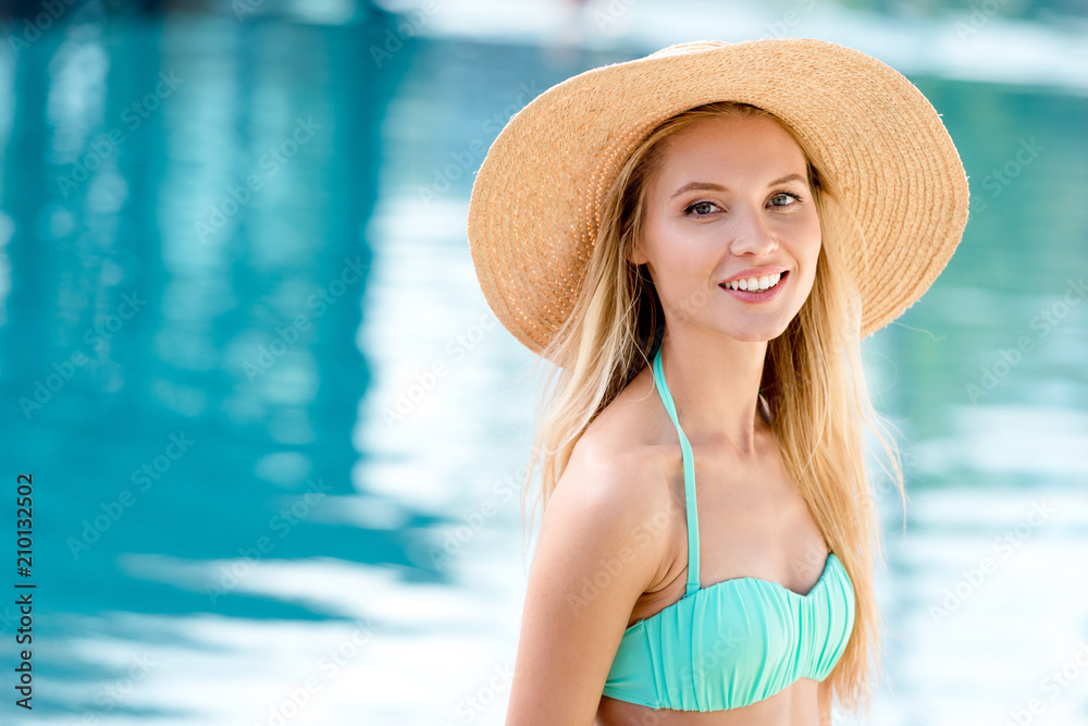 attractive young woman in straw hat and bikini looking at camera at poolside