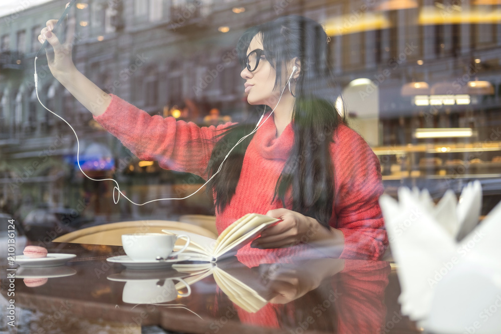 Cute brunette plus size woman working on a distance (freelance), sitting in cafe, drinking coffee and eating macaron. She wearing red sweater, listen music and make selfie on her phone