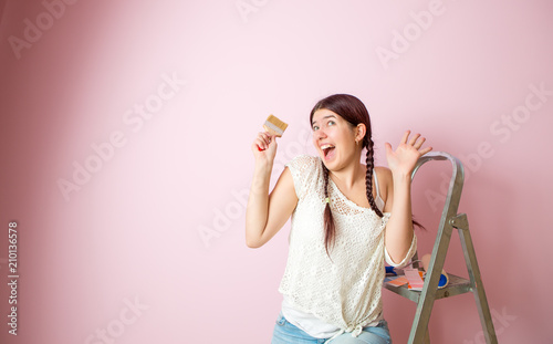 Photo of cheerful woman with brush next to stepladder and roller against blank pink wall.