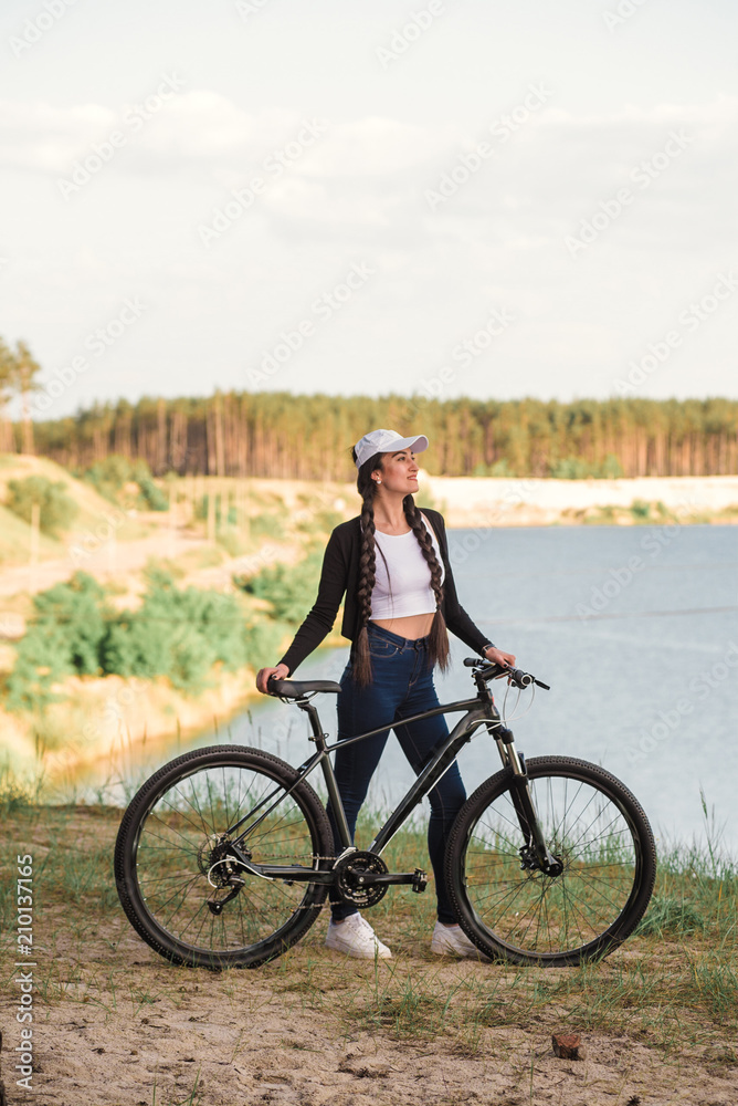 The beautiful girl is standing by bicycle. Senior woman riding a bike beside a lake. A female biker take a rest after biking The beautiful girl is standing by bicycle. Senior woman riding a bike beside a lake. A female biker take a rest after biking