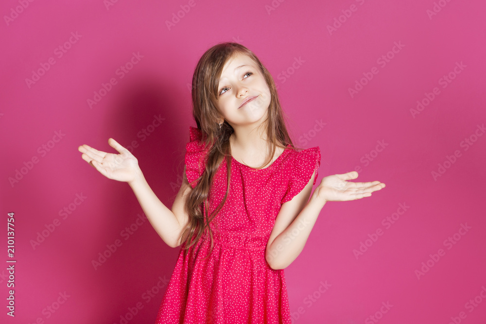 Little 8 years old girl make some emotional gesture with her hands on a pink neutral background. She has long brunette hair and wear red summer dress. Funny expression on her face