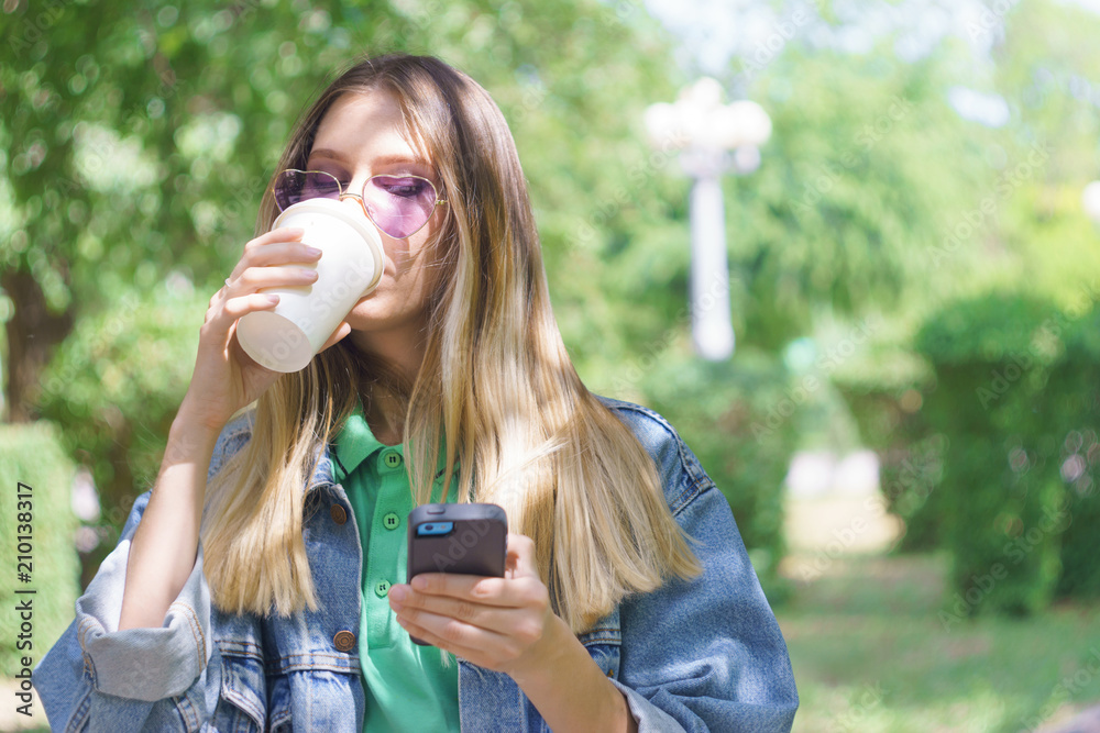 Attractive young girl is smiling and standing with a mug of coffee and a smartphone. Fashionable hipster model in trendy heart-shaped sunglasses. Concept of messaging and texting in the park Attractive young girl is smiling and standing with a mug of coffee and a smartphone. Fashionable hipster model in trendy heart-shaped sunglasses. Concept of messaging and texting in the park
