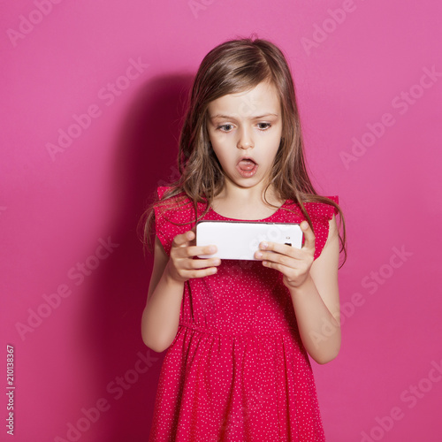 Little 8 years old girl make some emotional gesture with her hands on a pink neutral background. She has long brunette hair and wear red summer dress. Funny expression on her face