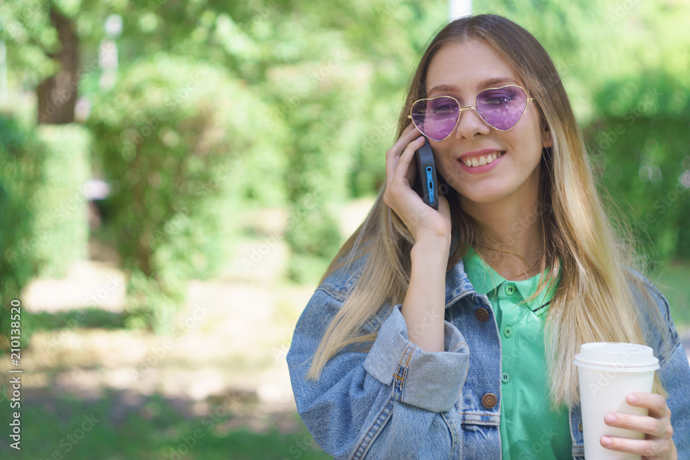 Attractive young girl is smiling and standing with a mug of coffee and a smartphone. Fashionable hipster model in trendy heart-shaped sunglasses. Concept of messaging and texting in the park