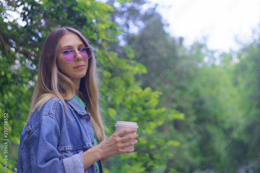 Cheerful young girl holding a cup with coffee. Fashionable smiling hipster model in trendy heart-shaped sunglasses. Face close up Cheerful young girl holding a cup with coffee. Fashionable smiling hipster model in trendy heart-shaped sunglasses. Face close up