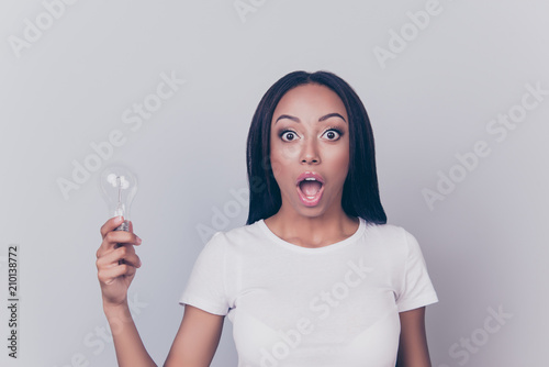 Incredible! Portrait of beautiful amazed astonished shocked surprised clever intelligent smart pretty afro brunette woman holding bulb in hand big eyes tanned bronze skin isolated on gray background photo