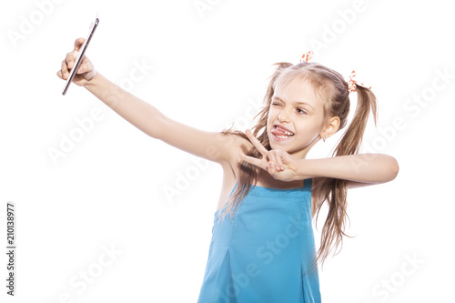 Young seven years old brunette girl in blue dress on a white isolated background. She makes selfie on her mobile phone, fun and smile emotions on her face