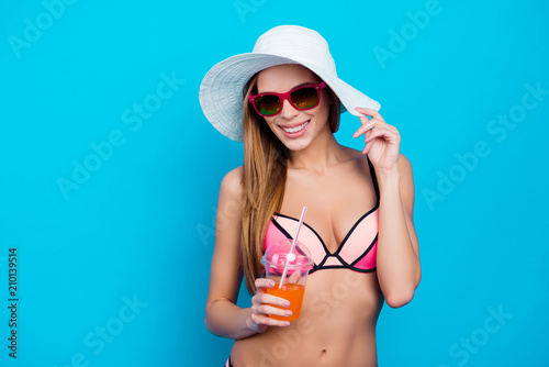 Portrait of cheerful positive girl in summer glasses swim wear holding hat and alcohol beverage with tubule isolated over blue background. Travel trip joutney tourism concept